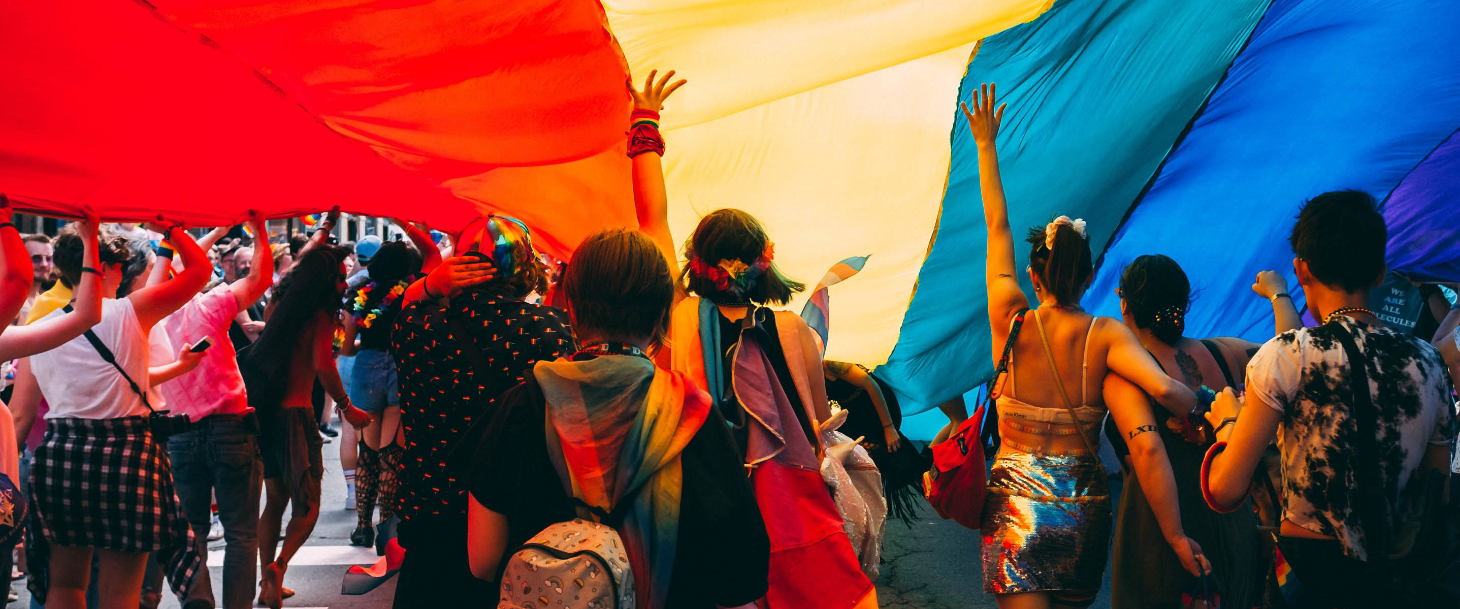 image of pride parade with attendees under a rainbow flag with arms upraised